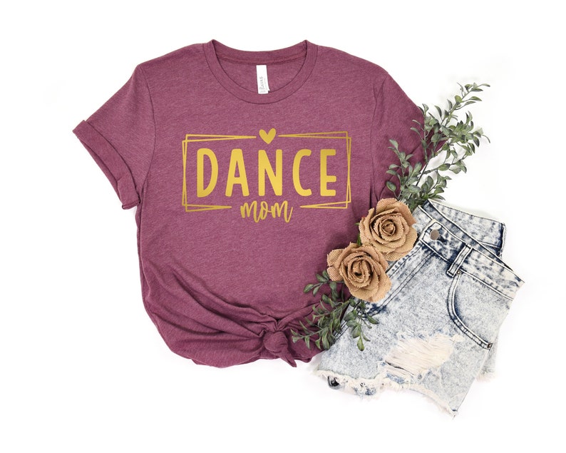 May include: A maroon t-shirt with the words "DANCE mom" in gold lettering inside a gold rectangle. The shirt is styled with denim shorts, faux flowers, and greenery on a white background.