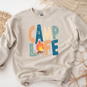 K&ouml;nnte beinhalten: Ein beiger Pullover mit dem Text "Camp Life" in bunten, abgenutzten Buchstaben. Ein Lagerfeuer mit orangefarbenen Flammen befindet sich mittig zwischen den W&ouml;rtern.