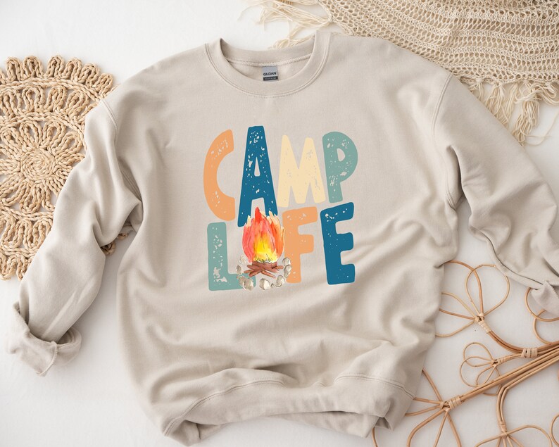 K&ouml;nnte beinhalten: Ein beiger Pullover mit dem Text "Camp Life" in bunten, abgenutzten Buchstaben. Ein Lagerfeuer mit orangefarbenen Flammen befindet sich mittig zwischen den W&ouml;rtern.
