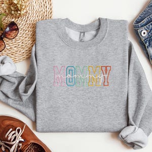 Pode incluir: Camisola cinzenta com a palavra "MOMMY" em cores do arco-&iacute;ris com uma fonte de escrita cursiva por baixo.