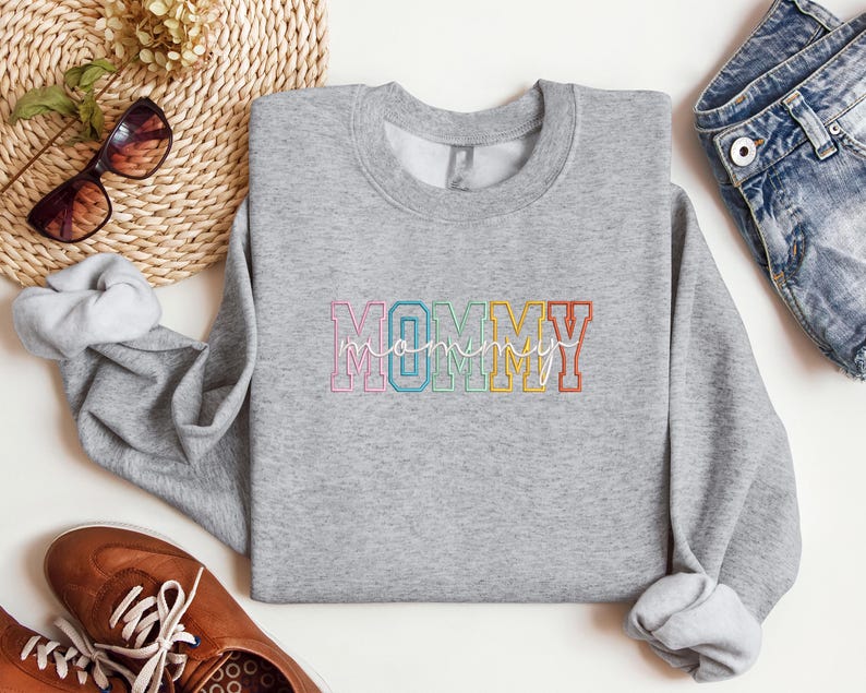 Pode incluir: Camisola cinzenta com a palavra "MOMMY" em cores do arco-&iacute;ris com uma fonte de escrita cursiva por baixo.