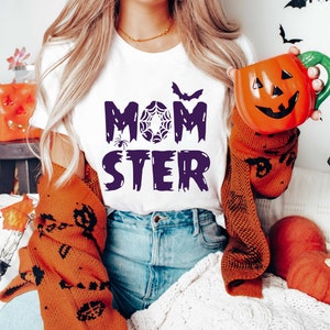 K&ouml;nnte beinhalten: Ein wei&szlig;es T-Shirt mit dem Text "MOM STER" in Lila, mit einer Spinnen- und Spinnennetzgrafik. Das Shirt wird von einer Person mit langen blonden Haaren getragen, die eine K&uuml;rbis-f&ouml;rmige Tasse in der Hand h&auml;lt.