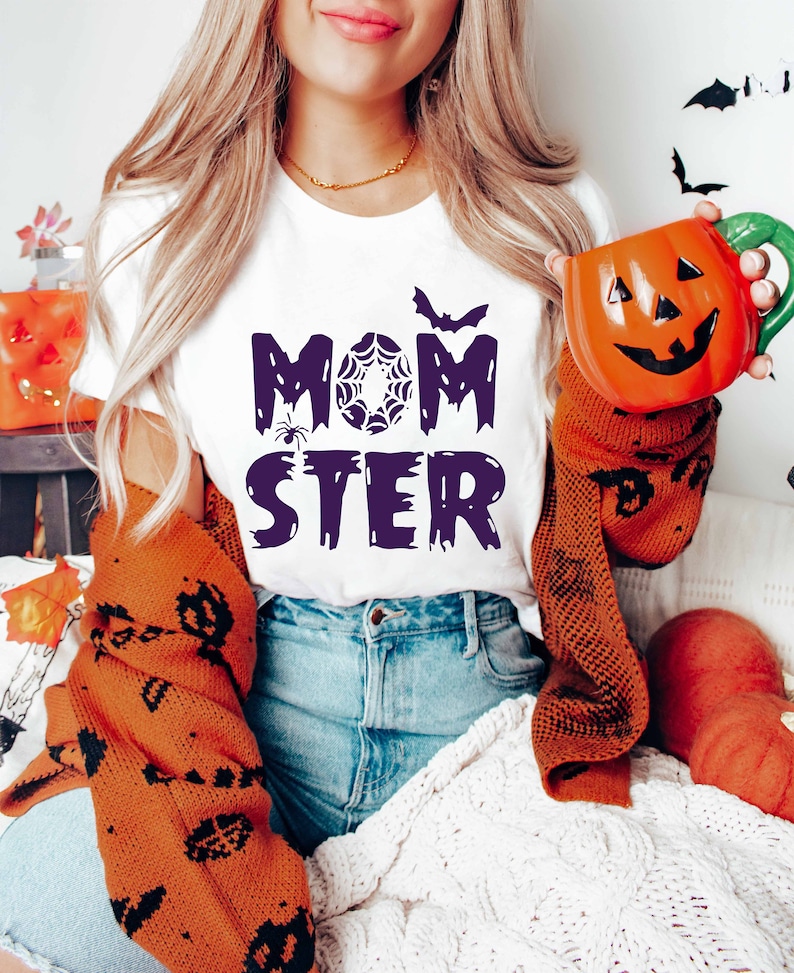 K&ouml;nnte beinhalten: Ein wei&szlig;es T-Shirt mit dem Text "MOM STER" in Lila, mit einer Spinnen- und Spinnennetzgrafik. Das Shirt wird von einer Person mit langen blonden Haaren getragen, die eine K&uuml;rbis-f&ouml;rmige Tasse in der Hand h&auml;lt.