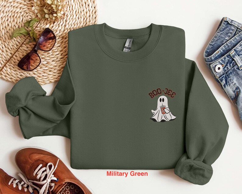 May include: A military green sweatshirt with a white ghost holding a cup of coffee and the text "Boo-Jee" on the front.