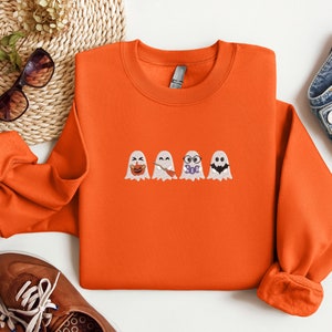 May include: Orange sweatshirt with a row of four embroidered ghosts. Each ghost has a different expression, including a pumpkin, a wand, glasses, and a bat.
