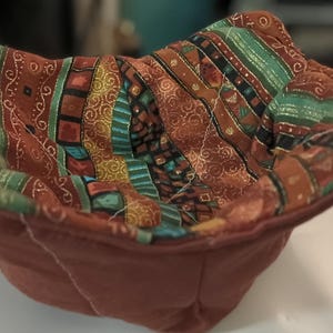 May include: A handmade, quilted bowl cozy in shades of brown, teal, and gold. The interior features a patterned fabric with stripes and geometric designs. The exterior is a solid, muted brown.