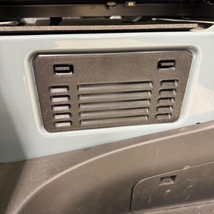 May include: A black plastic vent cover with horizontal slats and rectangular openings. The cover is mounted on a light blue surface, with a dark gray interior visible below. The design suggests automotive or vehicle interior components.