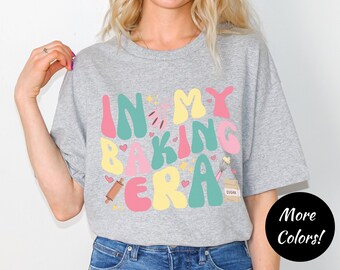 May include: Gray t-shirt with the text "In My Baking Era" in colorful, stylized letters. The text is surrounded by hearts, stars, and baking-themed illustrations.