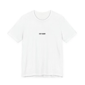 以下が含まれることがあります： 白のクルーネックTシャツ。胸の中央に黒字で「STAY SHARP.」とプリントされています。半袖で、クラシックでカジュアルなデザインで、普段使いに適しています。