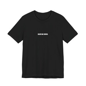 May include: Black crew neck t-shirt with the white text "DISCIPLINE DRIVEN" centered on the chest. The shirt has short sleeves and a classic fit, suitable for casual wear. The fabric appears to be a soft, comfortable material.