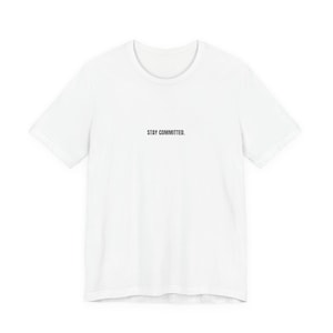May include: A white t-shirt with the text "STAY COMMITTED" printed in black, centered on the chest. The shirt has a crew neck and short sleeves. The fabric appears to be a soft, lightweight material.