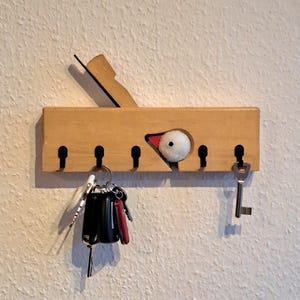 Könnte beinhalten: Schlüsselhalter aus Holz mit Vogeldesign. Der Schlüsselhalter ist aus hellfarbenem Holz gefertigt und zeigt einen geschnitzten Vogelkopf mit rotem Schnabel und weißem, flauschigem Körper. Mehrere Schlüssel hängen am Schlüsselhalter.