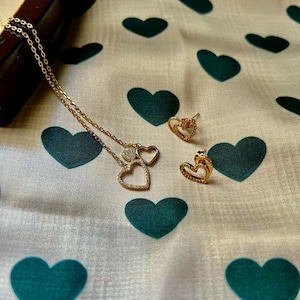 May include: A gold-colored necklace with heart-shaped pendants and matching heart-shaped stud earrings. The jewelry is displayed on a white fabric with a pattern of teal hearts.