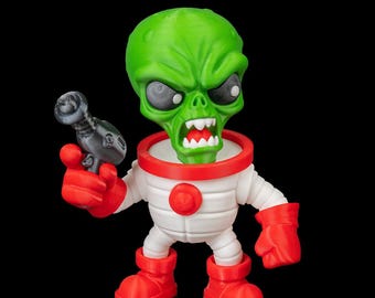 Puede incluir: Una figura de alienígena verde con una expresión enfadada, que lleva un traje espacial blanco con detalles rojos. La figura sostiene una pistola de rayos gris en su mano derecha y tiene guantes y botas rojas. La figura es un juguete o un objeto de colección.