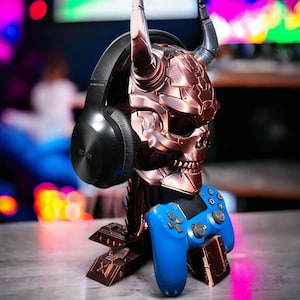 Puede incluir: Un soporte de color cobre en forma de cráneo con cuernos que sostiene un par de auriculares inalámbricos negros y un mando de PlayStation azul.