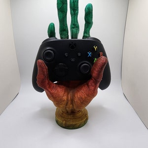 Può includere: Una mano stampata in 3D che tiene un controller per videogiochi nero. La mano è dipinta in tonalità di arancione, rosso e giallo. Le dita sono verdi.