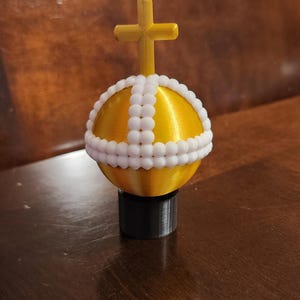 May include: A gold orb with a white beaded band and a gold cross on top. The orb is sitting on a black base.