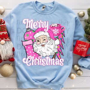 Puede incluir: Una sudadera azul claro con un diseño navideño rosa y verde que representa a Papá Noel sosteniendo una media y una taza de chocolate caliente. El texto "Merry Christmas" está escrito en letras rosas.