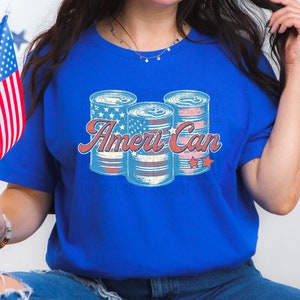 Puede incluir: Una camiseta azul con un gráfico de tres latas rojas, blancas y azules con estrellas. El texto "Ameri Can" está impreso en la camiseta.