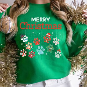 Puede incluir: Suéter navideño verde con el texto "Feliz Navidad" y un diseño de huellas de patas con decoraciones navideñas colgando.