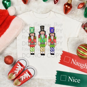 Könnte beinhalten: Weißes T-Shirt mit drei bunten Nussknacker-Illustrationen. Das Shirt trägt den Text "Neon Flamingo Shop Do Not Copy". Es gibt zwei rot-grüne Bänder mit dem Text "Naughty" und "Nice" darauf.
