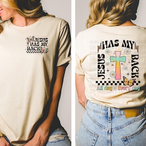 May include: A beige t-shirt with a graphic design on the front and back. The front design says "Jesus Has My Back" with butterflies and a checkered pattern. The back design says "Jesus Has My Back" with a cross, butterflies, flowers, and the text "All day Every day".