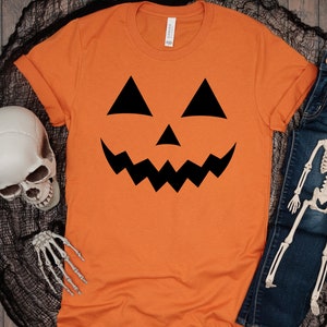 以下が含まれることがあります： オレンジ色のTシャツに、黒いジャック・オ・ランタンの顔のグラフィックがプリントされています。このTシャツは、ハロウィンのコスチュームとして人気があります。