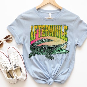 May include: Light blue t-shirt with a graphic of an alligator and the text "Afterwhile" in a retro font. The alligator is green and has a yellow, orange, and red sunset behind it.