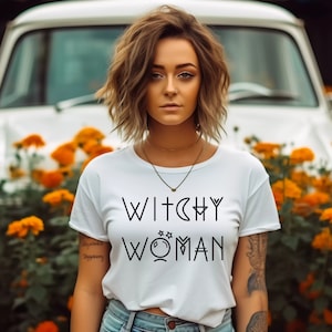 May include: A white t-shirt with the text "Witchy Woman" in black lettering. The text includes a crescent moon, a star, and a circle with a star inside.