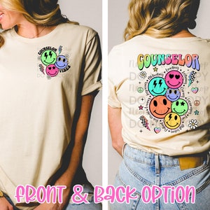 May include: A beige t-shirt with a colourful graphic design on the front that says "COUNSELOR VIBES". The back of the shirt has a larger graphic design with the word "COUNSELOR" and several smiley faces with different expressions and words like "leading", "motivating", "listening", "caring", "friendly", "helpful", and "encouraging".