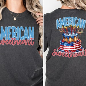 May include: A dark gray t-shirt with the text "American Sweetheart" in blue and red lettering. The back of the shirt features a red, white, and blue cake with candles and fireworks.