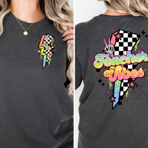 May include: Two images of a dark grey t-shirt with a graphic design. The design on the left is a rainbow coloured lightning bolt with a chequered pattern. The design on the right is a rainbow coloured lightning bolt with a chequered pattern and the text "Teacher Vibes".