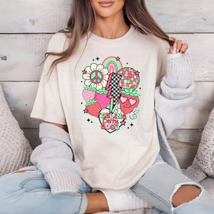 Può includere: Una maglietta bianca a maniche lunghe con una stampa grafica di una fragola, un segno di pace, un arcobaleno, un fulmine e una palla da discoteca. Il testo "Feeling Berry Good" è stampato sulla maglietta.