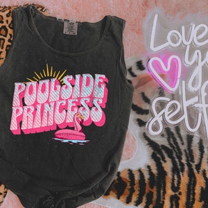 May include: Black tank top with a pink graphic that says "Poolside Princess" with a pink flamingo pool float. The graphic has a retro style.