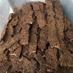 May include: A close-up shot of a clear plastic container filled with strips of dark brown beef jerky. The jerky pieces are stacked and layered, showing a textured surface. The jerky appears to be dried and seasoned.