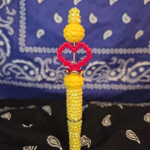 May include: A pen featuring a yellow, textured, iridescent barrel and a matching yellow bead topper. The pen includes a bright red, pixelated heart-shaped charm positioned between the yellow beads, creating a distinct and colorful writing instrument.