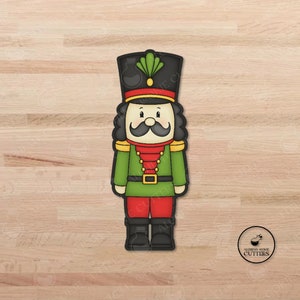 May include: A green, red, and black cookie cutter shaped like a traditional Christmas nutcracker. The nutcracker has a black hat with a green feather, a white face with a black mustache, and a red sash.