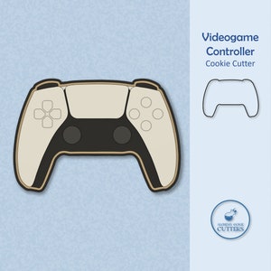 May include: A white and brown cookie cutter shaped like a video game controller. The controller has two black buttons and a black directional pad. The cookie cutter is for making cookies in the shape of a video game controller.