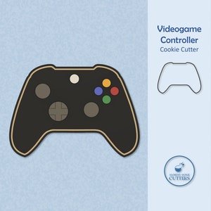 May include: Black and gold video game controller cookie cutter. The cutter has a white button, a black directional pad, and four colored buttons. The cutter is shown against a light blue background.