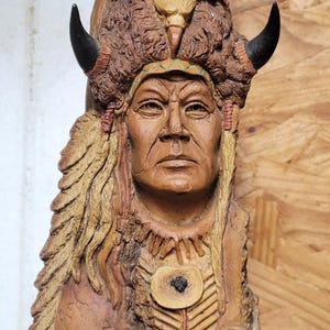May include: A detailed wood carving of a Native American chief. The sculpture features a headdress with horns, a stern facial expression, and intricate details. The carving is in shades of brown and tan.