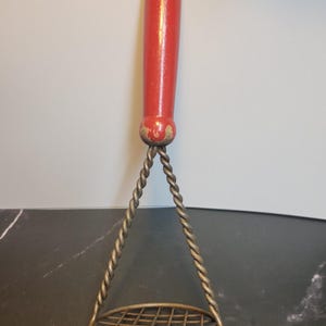 May include: A vintage potato masher featuring a cylindrical red painted wooden handle with visible wear at the base. The masher has a metallic, twisted wire frame that connects to a circular, grid-patterned mashing plate at the bottom.