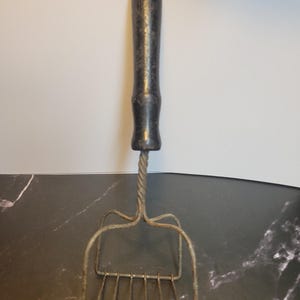 May include: A vintage potato masher featuring a dark, weathered wooden handle attached to a twisted metal shaft. The mashing head is constructed from a single piece of bent wire, forming a rectangular grid pattern designed for crushing food items.