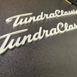 May include: Two silver-colored, three-dimensional script emblems featuring the text TundraClassic. Each emblem has a metallic, brushed finish with raised lettering and a cursive font style, positioned diagonally across the frame.