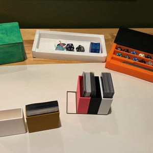 Könnte beinhalten: Eine Auswahl an Tabletop-Gaming-Zubehör, darunter eine grüne Würfelbox, weiße und orangefarbene Tabletts und Kartenhalter in verschiedenen Farben. Mehrere Würfel werden in den Tabletts ausgestellt, und Kartenstapel sind in den Haltern sichtbar.