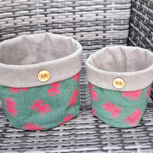 May include: Two fabric storage baskets. The baskets have a green base with a red dragon pattern and a grey linen top. Each basket has a wooden button detail. The baskets are on a woven surface.