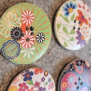 May include: Four decorative wooden buttons with floral patterns. The buttons are in various colours, including green, white, red, blue, and purple. Each button has a unique floral design and several holes for sewing. The buttons are approximately 3 cm in diameter.