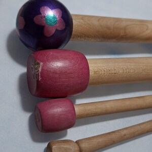 May include: A selection of wooden tools with different head designs. The top tool features a purple ball head decorated with pink and green floral patterns. Other tools have pink and natural wood heads. The handles are made of natural wood.