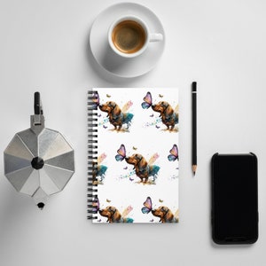 May include: A white spiral bound notebook with a repeating pattern of a brown and black dachshund dog with a butterfly in its mouth. The butterfly is purple, pink, and blue. The background is white with watercolor splatters.