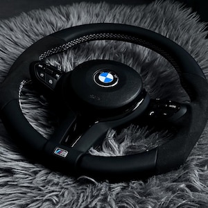 May include: A black BMW steering wheel with a suede grip and white stitching. The center features the BMW logo in blue and white. The wheel has integrated controls and the M logo.