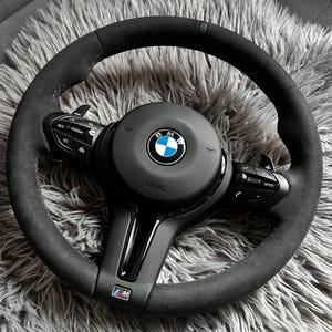 May include: A black BMW steering wheel with a suede-like grip. The center features the BMW logo and the word "AIRBAG". The wheel has integrated controls and paddle shifters. The bottom of the wheel has the M logo.